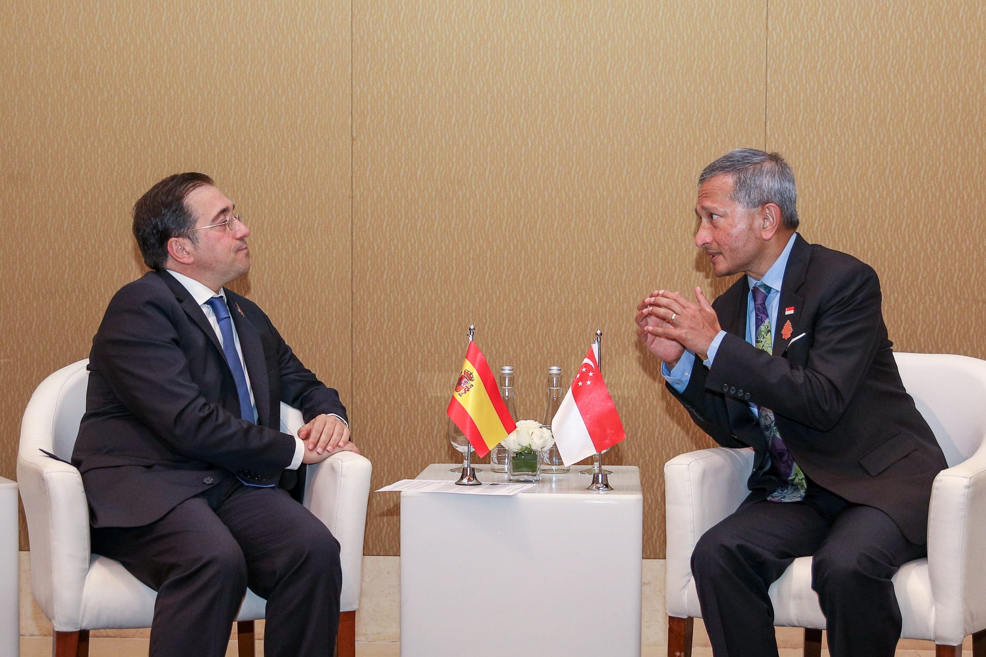 Two men in suits seated facing each other with Singapore and Spain flags on a table.